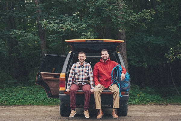 two men sitting in the open boot of a car in hiking gear with a backpack